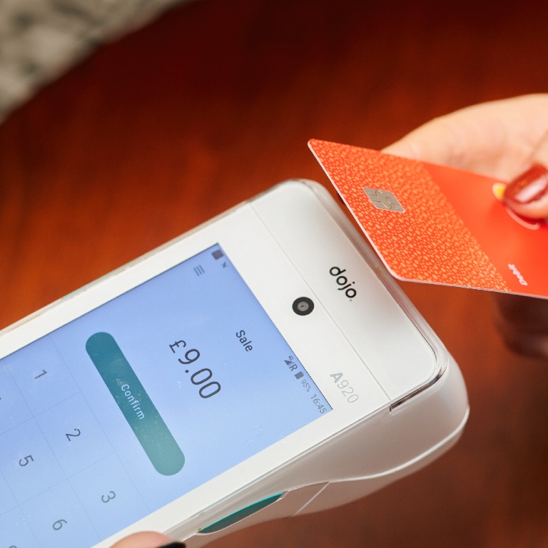 Hand tapping a red bank card on a Dojo payment terminal showing a GBP 9.00 sale, illustrating cyber risk for pub operators.